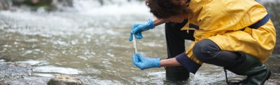Registered Environmental Technician in a yellow coat taking a sample of water from a river society for the environment