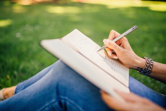 Close up of writing notes in a note book in the park with greenery in the background. Resting note pad on legs wearing jeans. EnvCast environmental podcast
